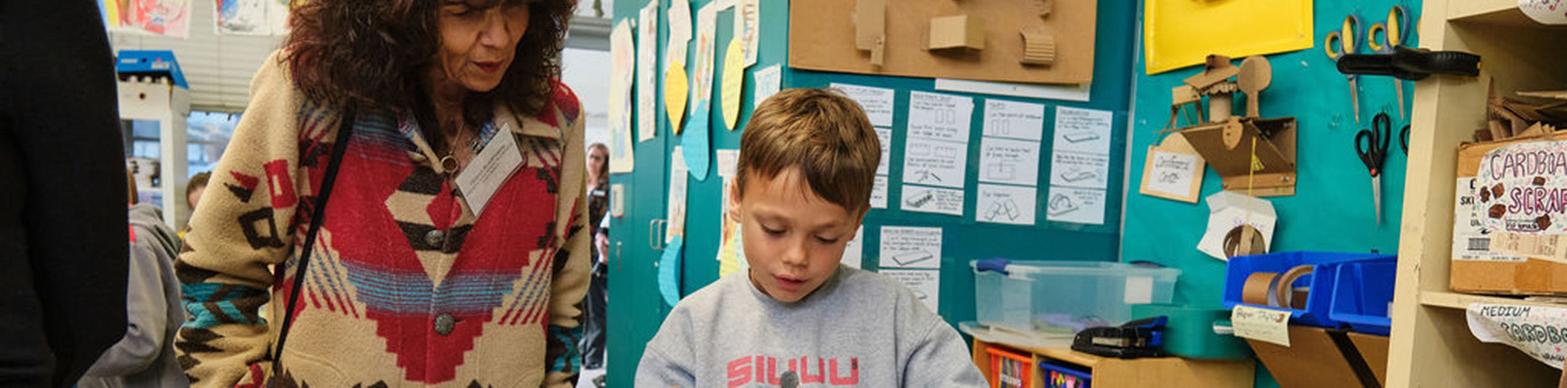 Older brown-skinned woman looks on while a young white boy creates a sculpture