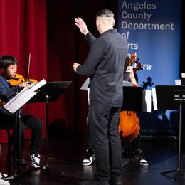 A conductor dressed in all black stands at the center of the stage, facing a row of elementary students dressed in navy T-shirts and dark pants. The students are performing on violin, viola, cello and double bass while referencing sheet music from the stands in front of them.